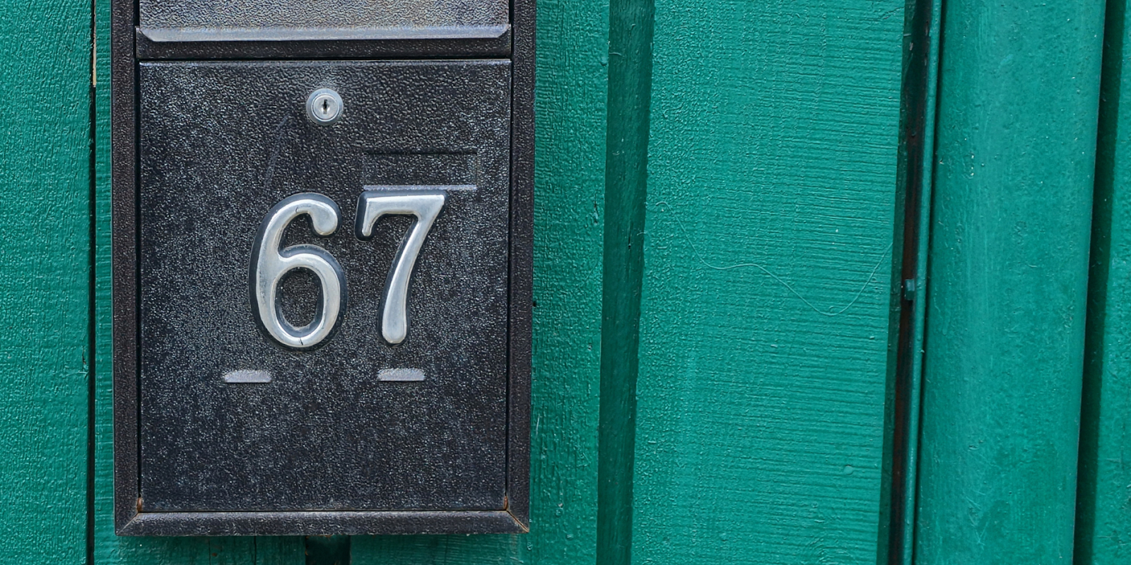 A traditional black mail box with number 67 on attached to a teal fence.